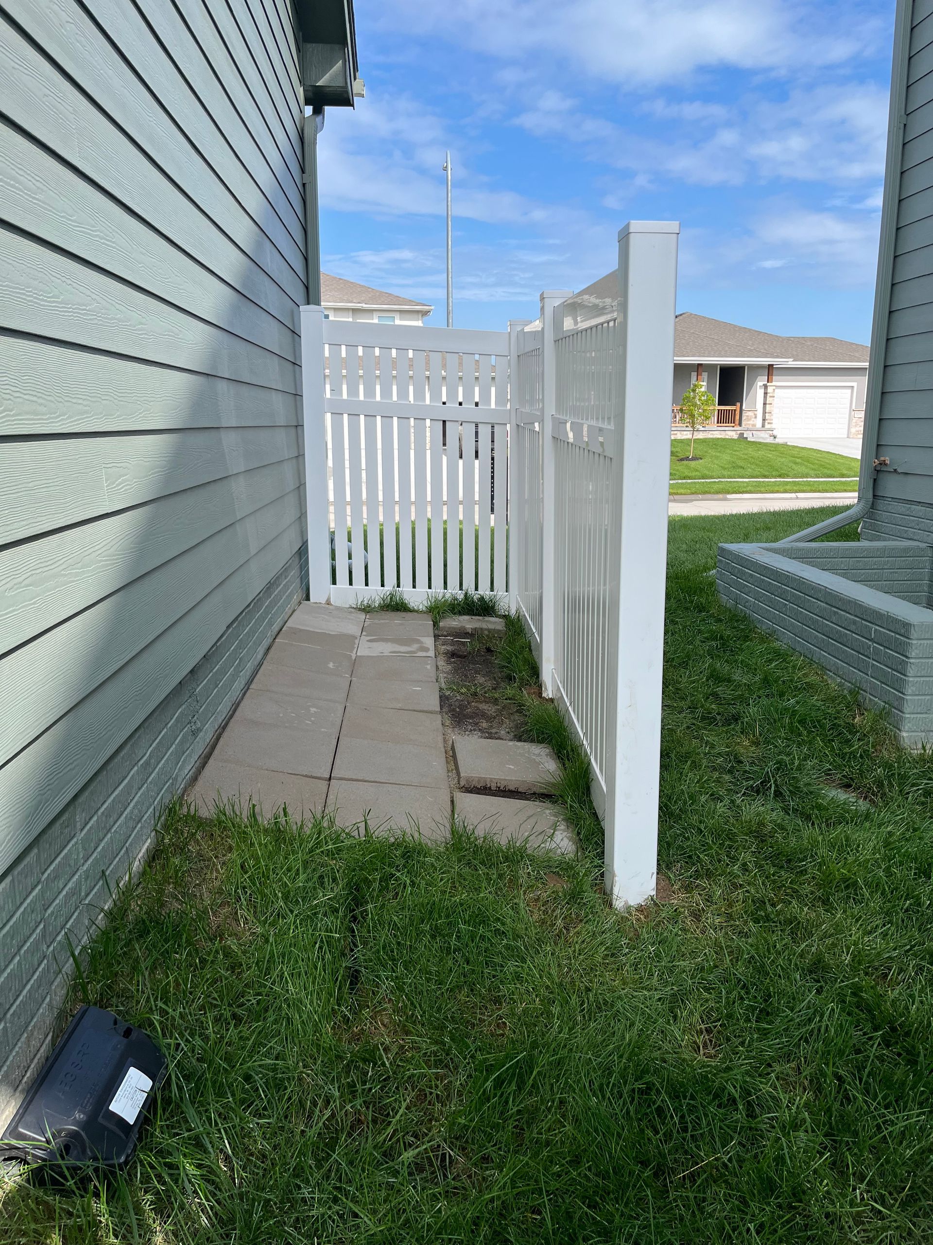 White fence section next to a house with concrete path and grass, sunny day.