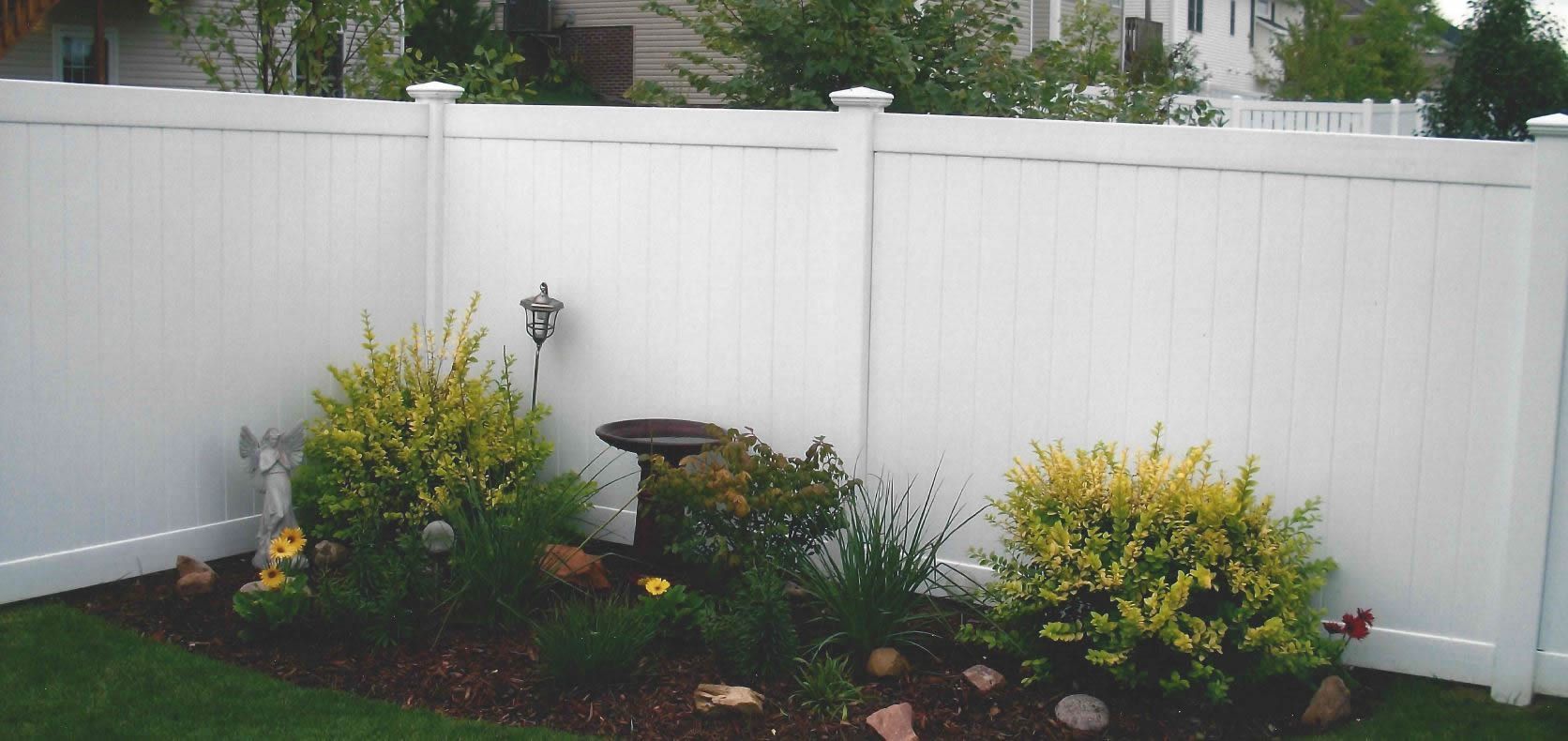 White fence encloses a garden bed with shrubs, flowers, and a birdbath on a green lawn.