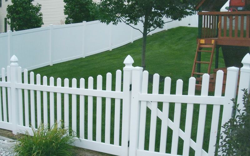 White picket fence with gate in front of a yard with a playset and a white fence in the background.