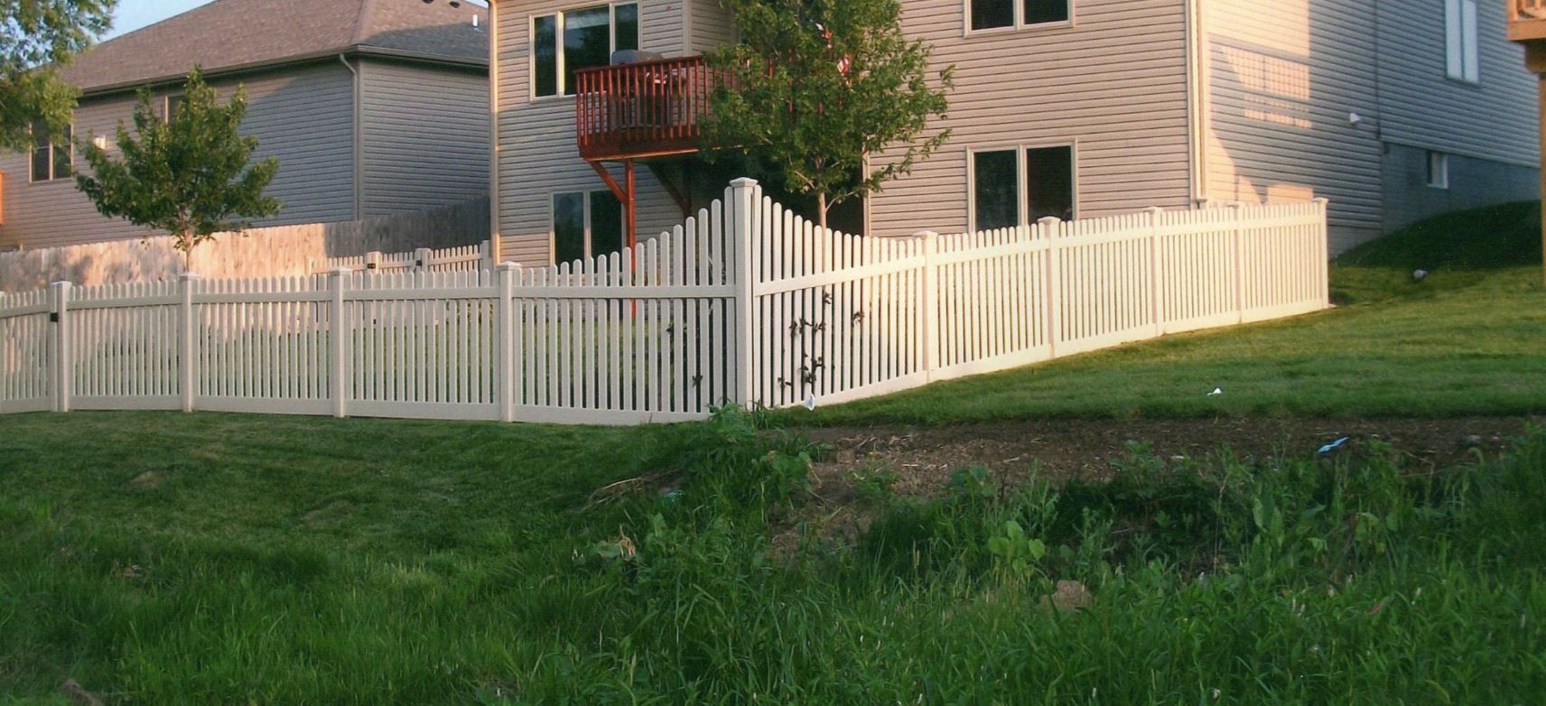 White picket fence in front of a house on a grassy hill, with bushes and trees surrounding.