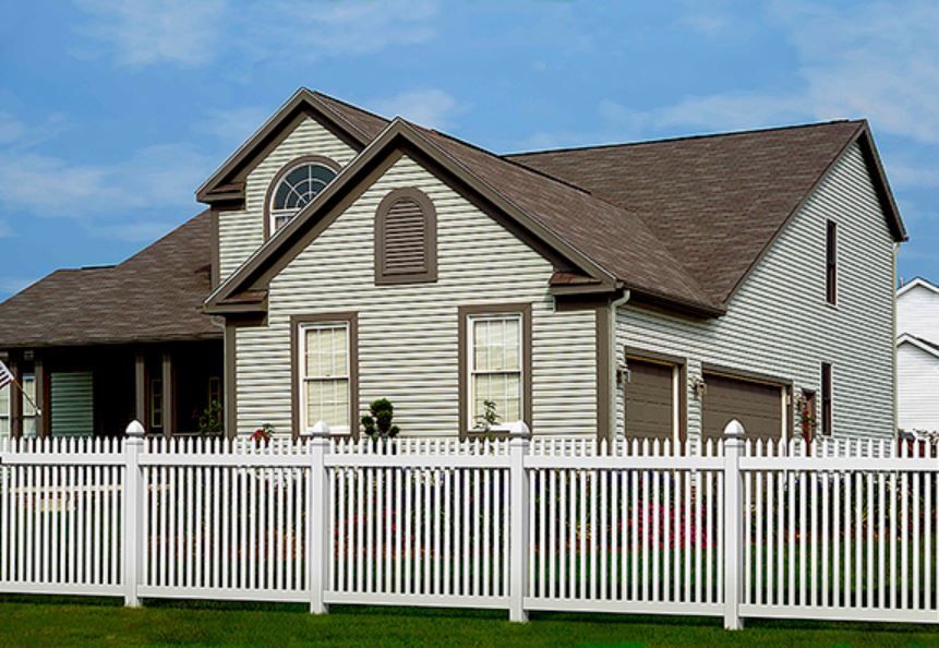 White picket fence in front of a light green house with brown roof and garage doors, blue sky.