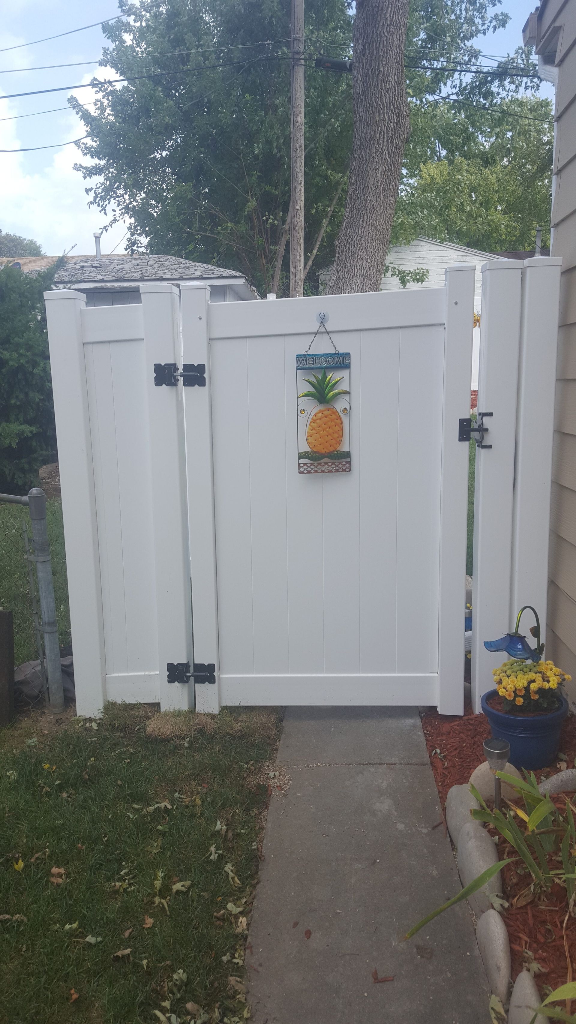 White vinyl gate with pineapple decor, leading to a yard with grass and flowers.