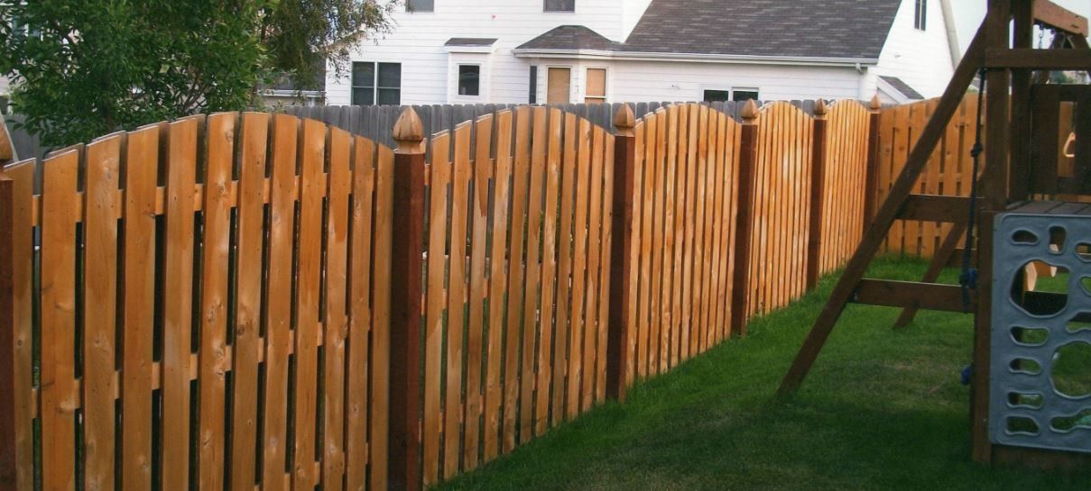 Wooden fence surrounding a grassy yard with a playground. A white house is in the background.