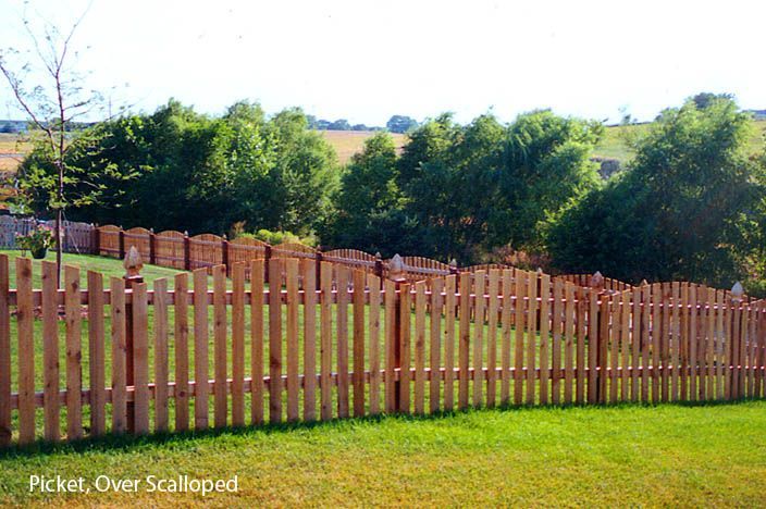 Wooden picket fence with scalloped top, enclosing a grassy yard with trees and rolling hills in the background.