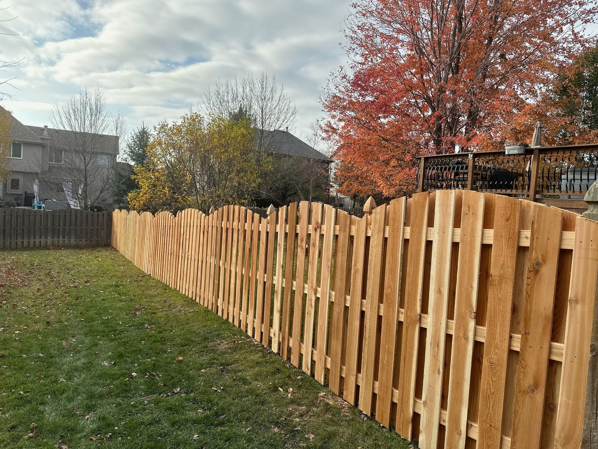 Wooden fence with a scalloped top, outlining a green lawn; autumn foliage in the background.