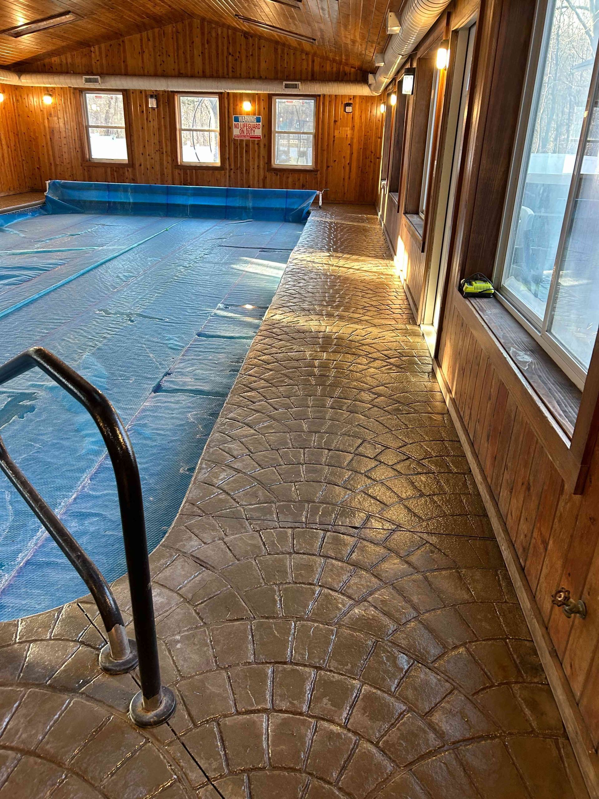 Indoor pool with dark brown brick-patterned walkway. The pool water is covered with a blue tarp.