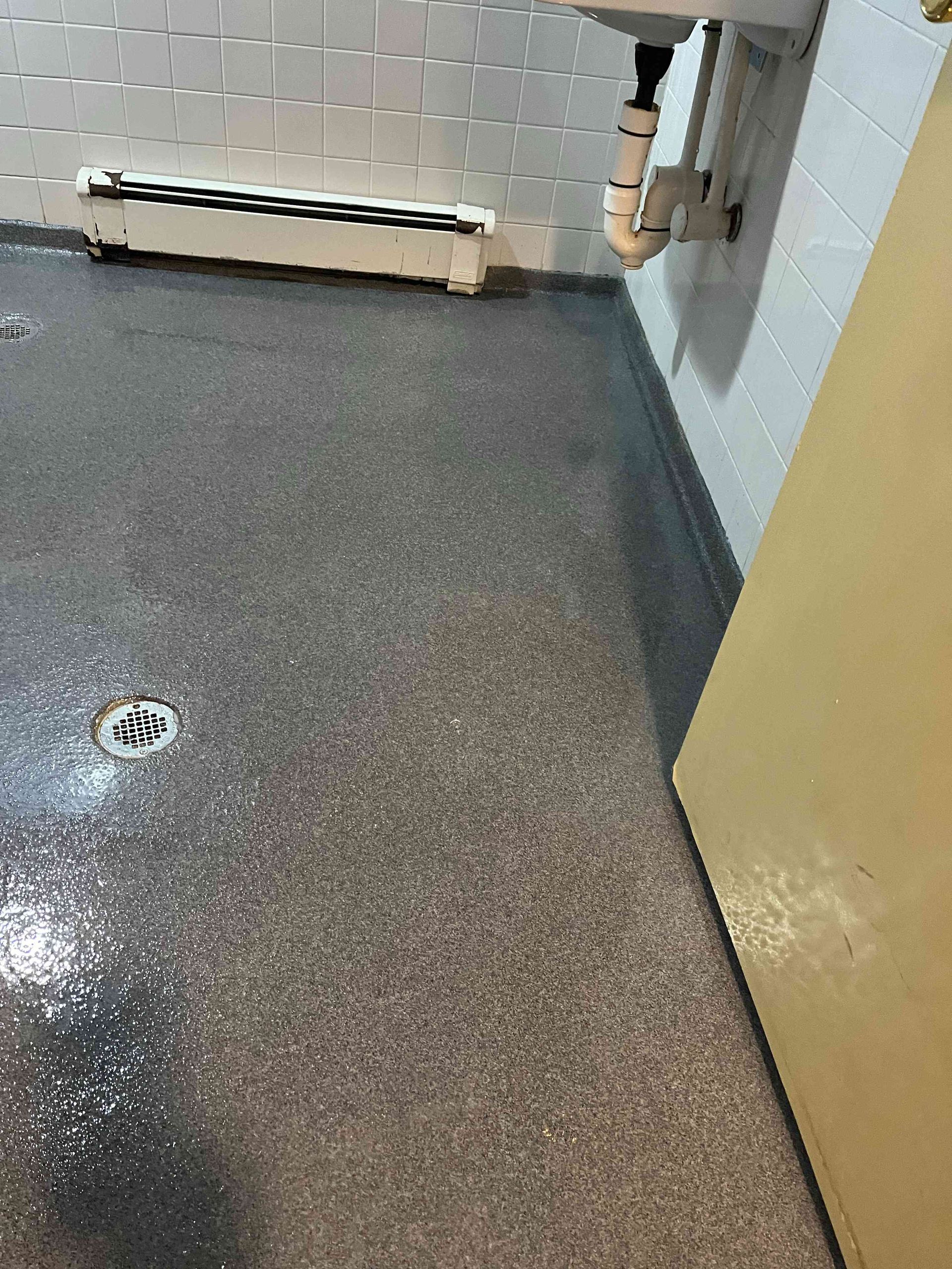 A wet, gray floor with a drain and a white sink against a tiled wall.