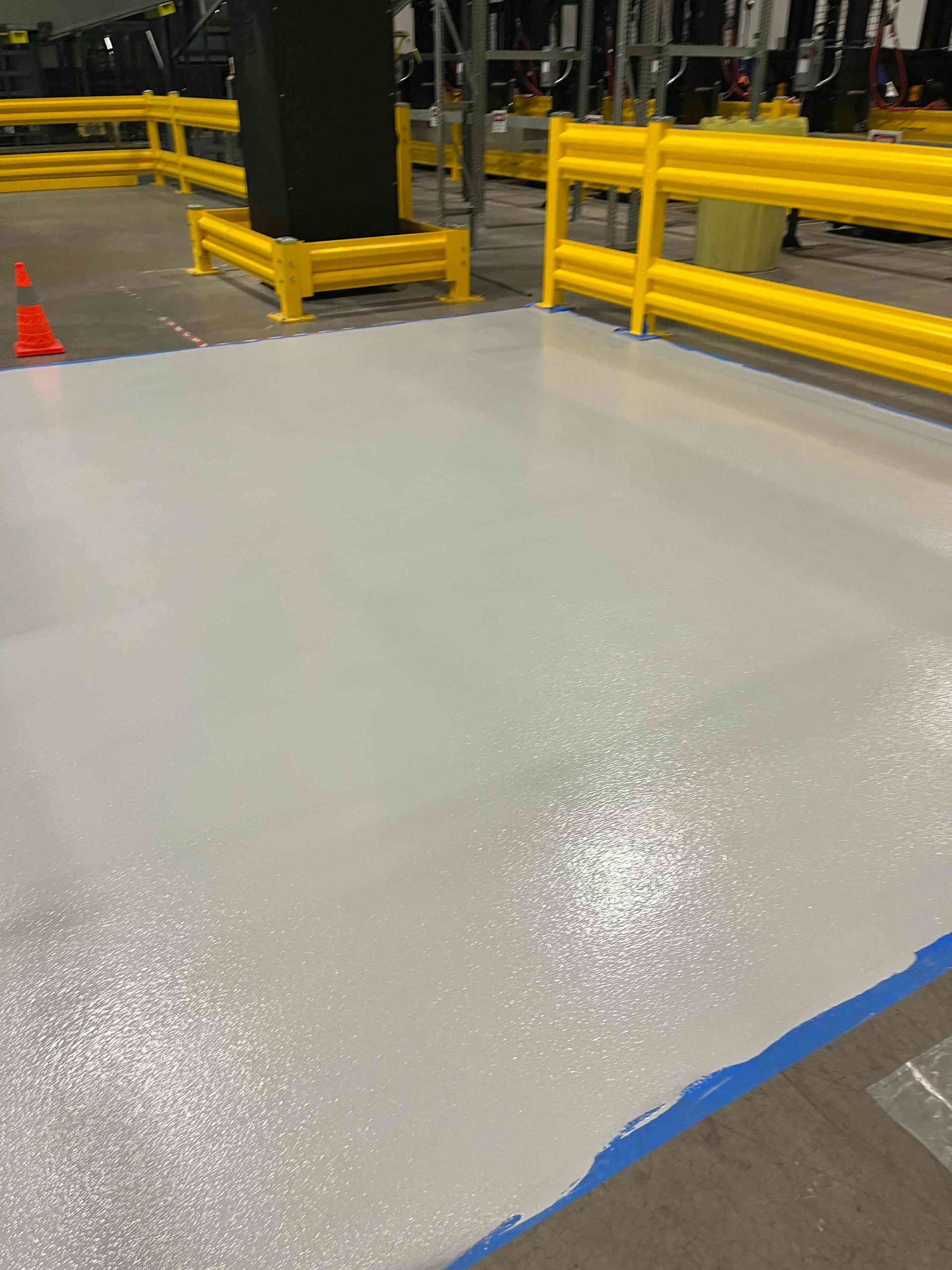 Newly painted light gray floor in industrial setting, bordered by yellow safety barriers.