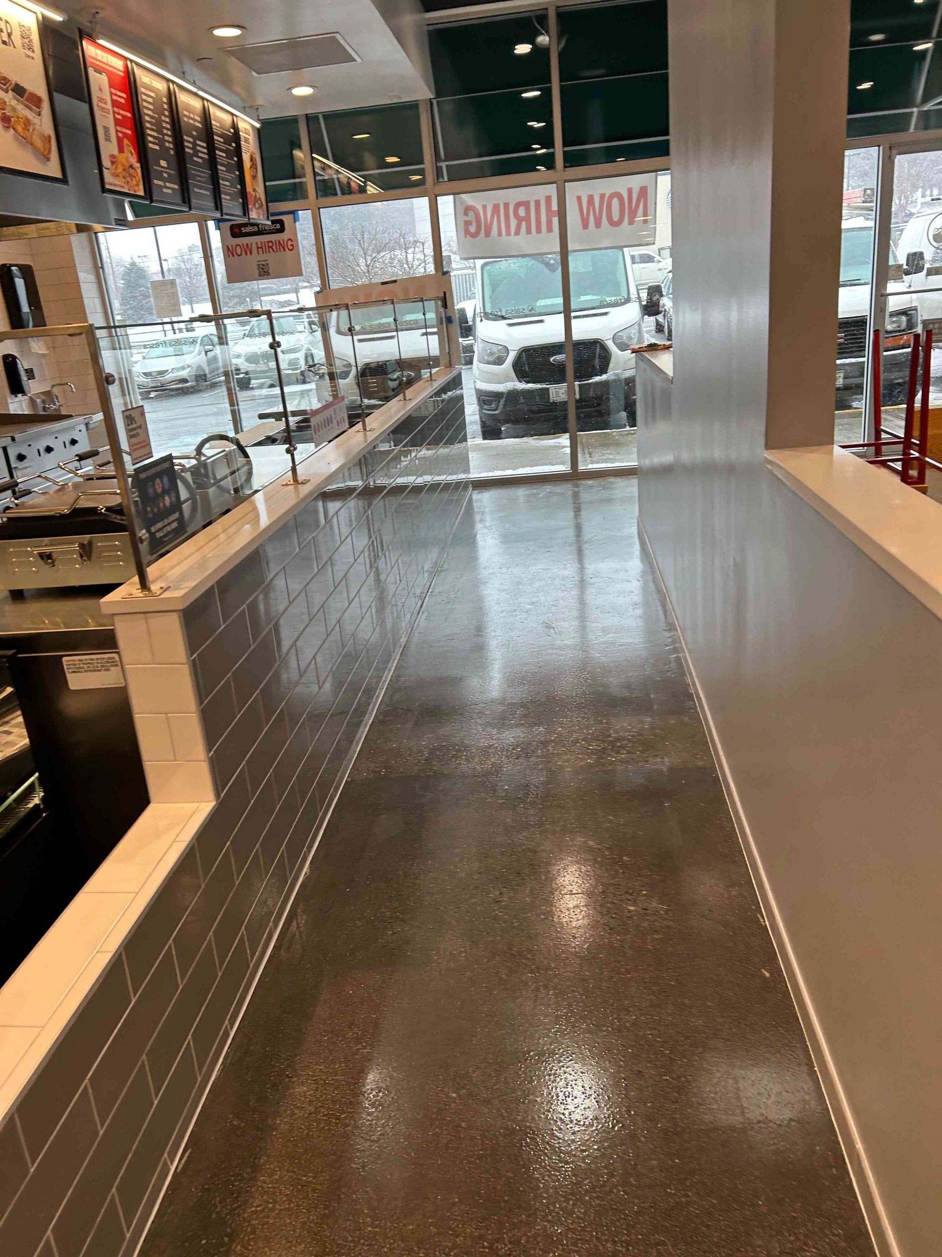 Interior view of a business with a wet, reflective concrete floor near large windows overlooking a parking lot with vehicles.