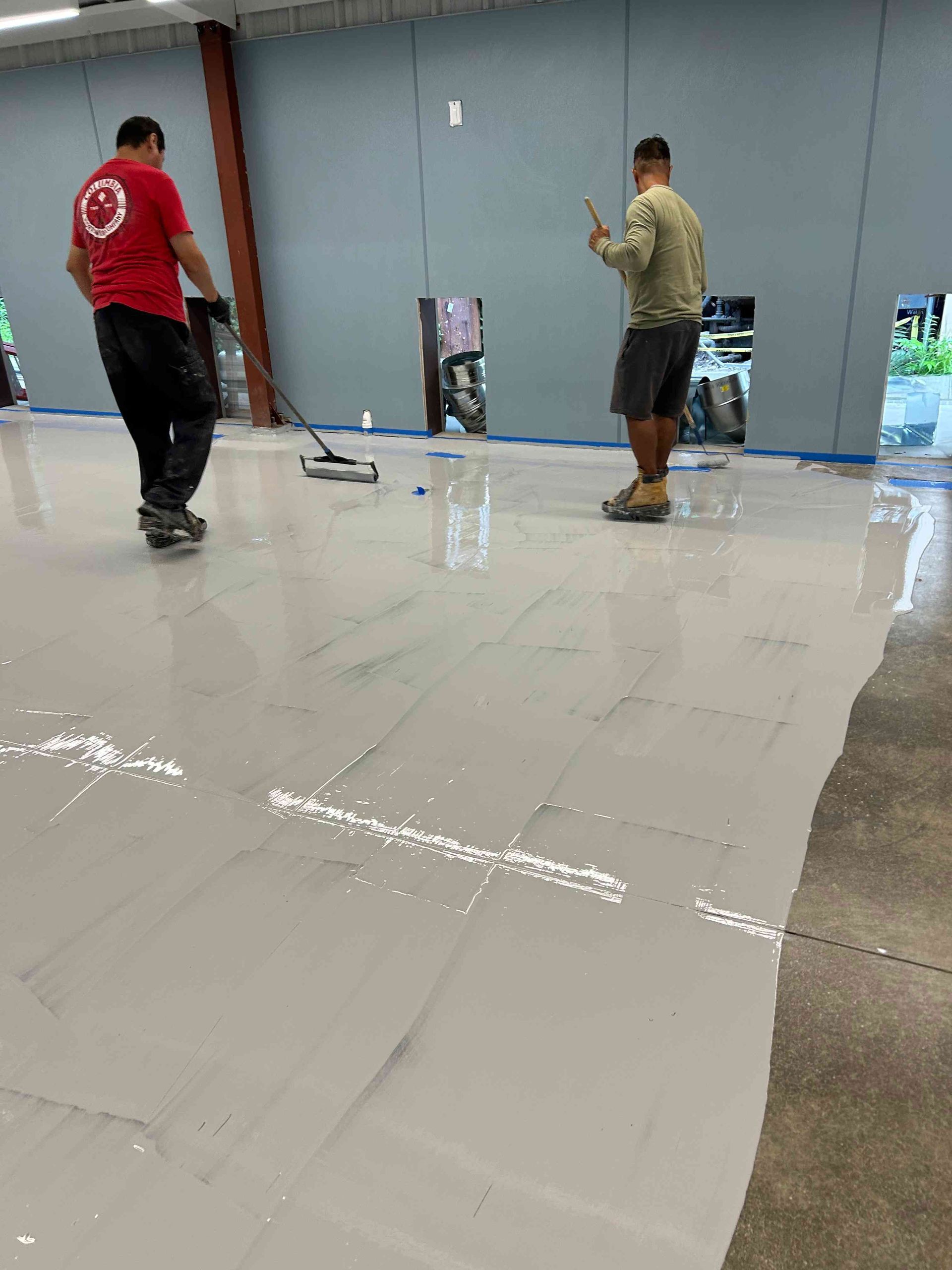 Two people applying gray epoxy flooring in a commercial space, using a roller and gesturing.