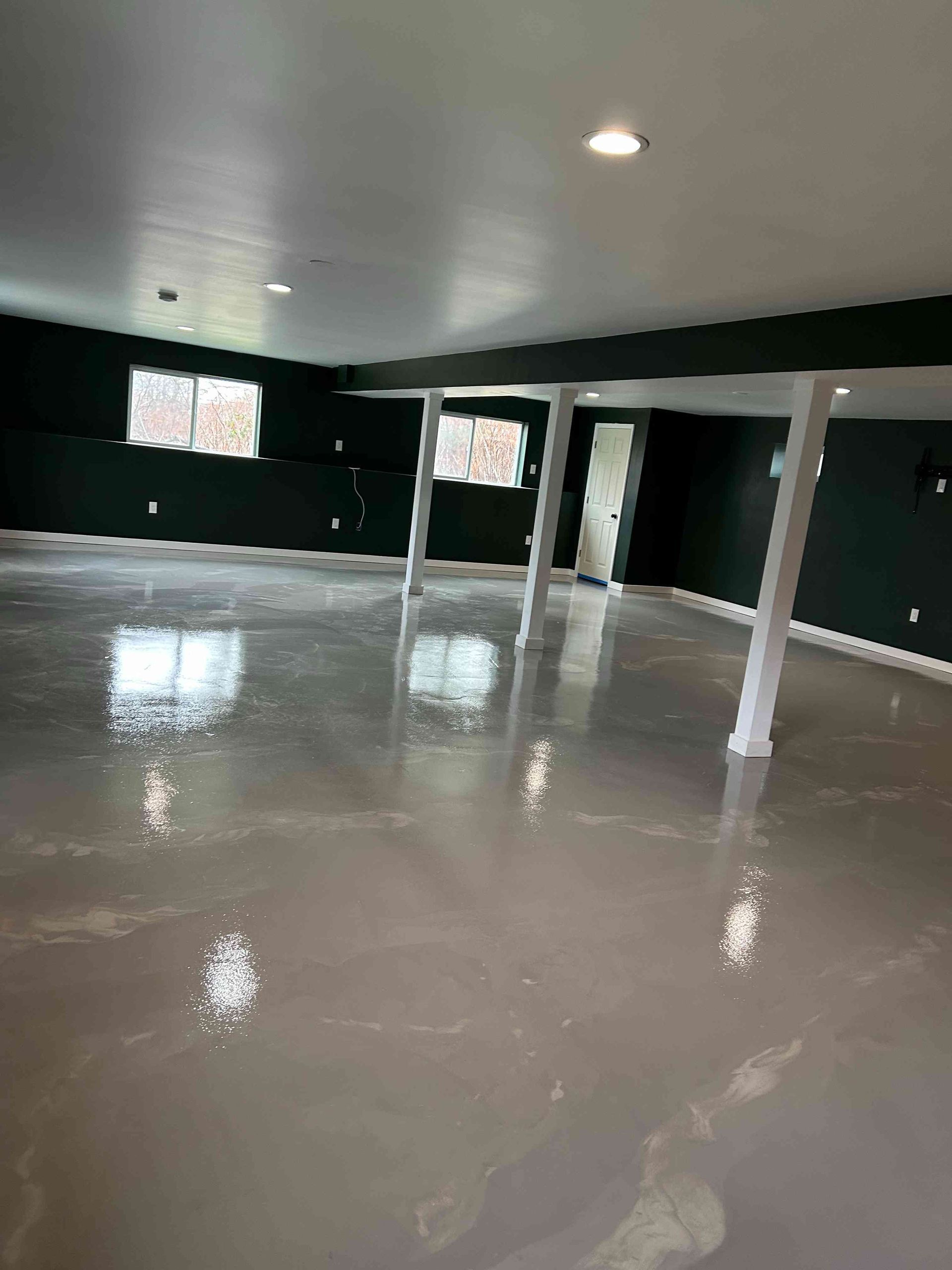 Empty basement with glossy, light-colored floor, dark green walls, white support beams, and white ceiling.