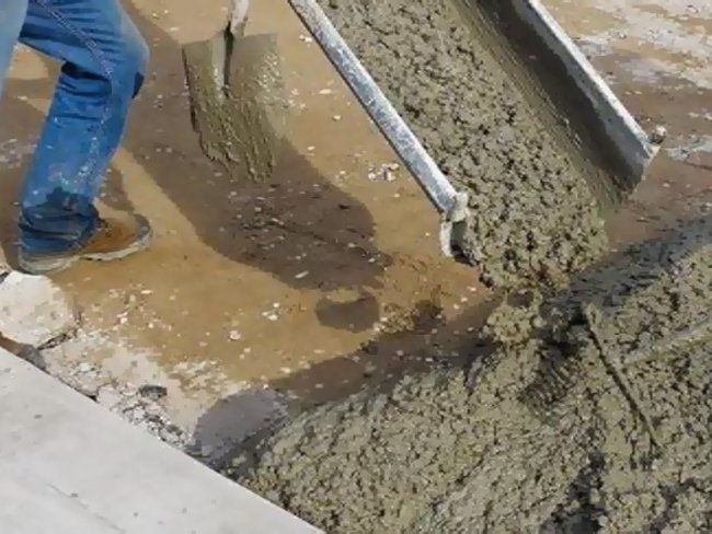 Person pouring wet cement with a shovel from a metal chute onto a paved surface.