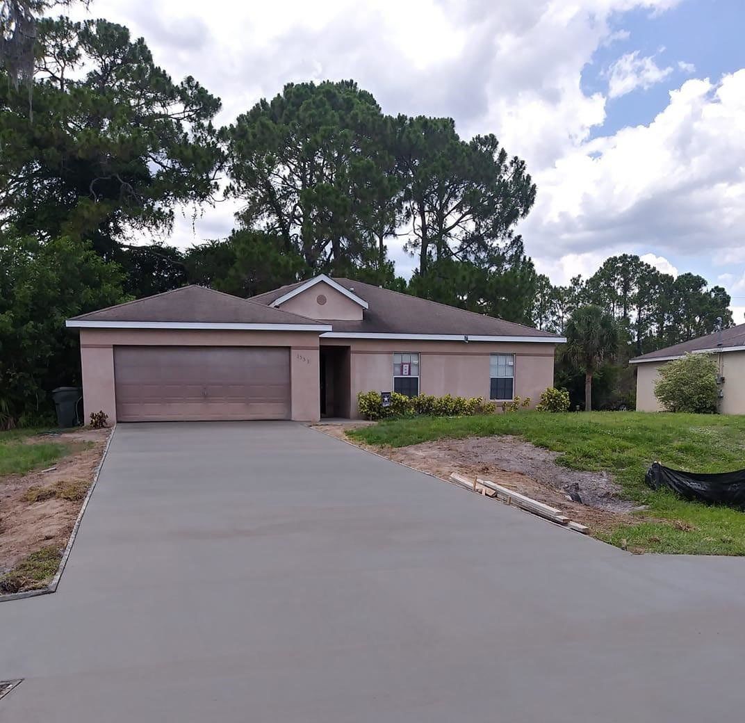 Tan house with a new concrete driveway, garage door, and landscaping, against a backdrop of trees and a cloudy sky.