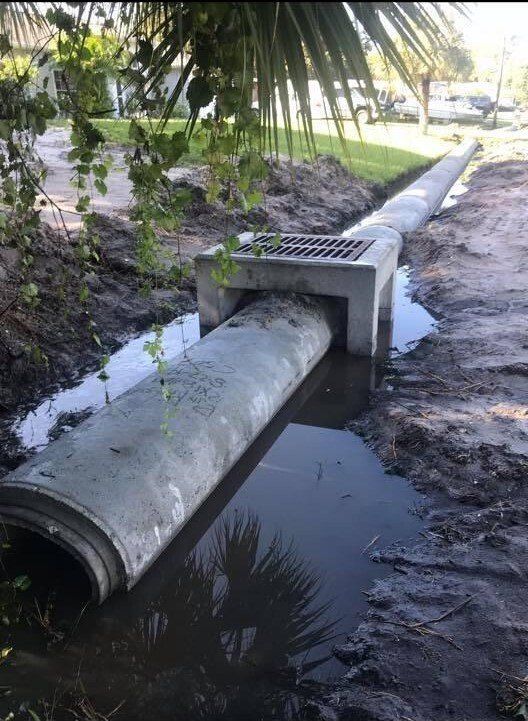 Concrete drainage pipe and grate along a street, with water visible in the ditch.
