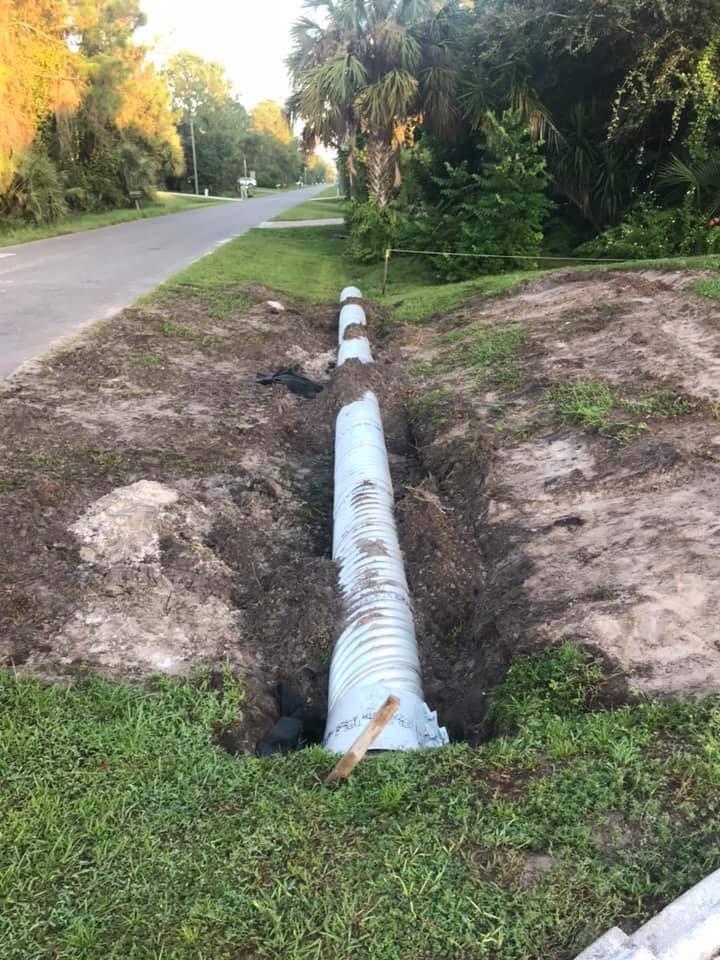 Ditch with exposed corrugated pipe next to a paved road, dirt, grass, and trees.