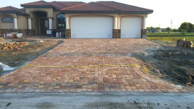Driveway of brick pavers leading to a beige house with two garage doors. Construction site.