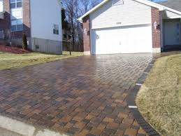 Brick driveway leading to a white garage door, adjacent to a grassy lawn and a house.