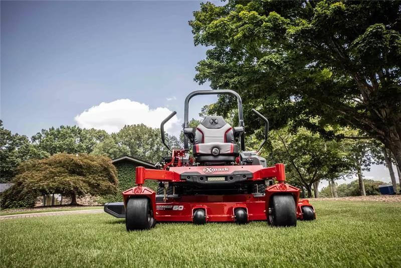 Red zero-turn lawnmower on green grass in front of trees under a blue sky.