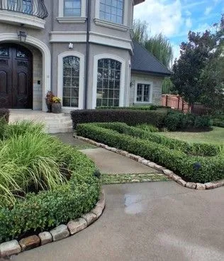 Stone-trimmed driveway and manicured shrubs lead to a gray stucco house with arched windows and a dark wood door.