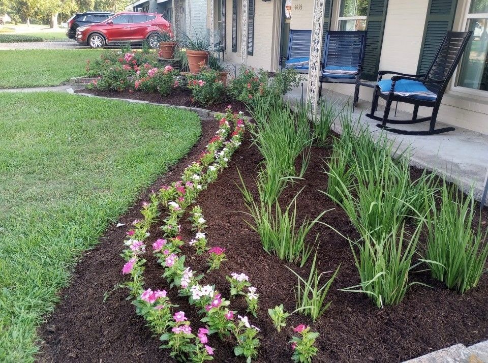 Well-landscaped front yard with flowers, mulch, and tall green plants. A red car is parked on the street.