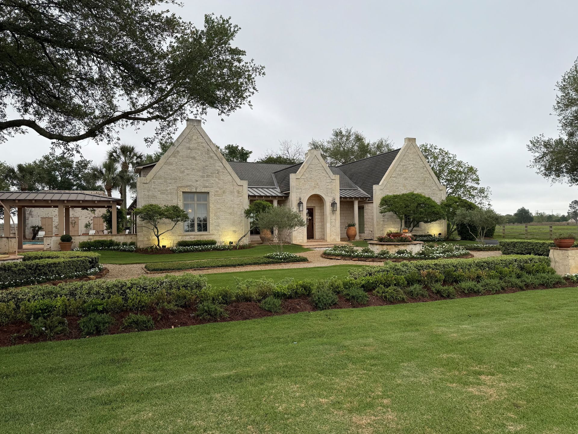 A tan stone house with a multi-gabled roof, framed by lush green lawns and hedges under a cloudy, overcast sky.