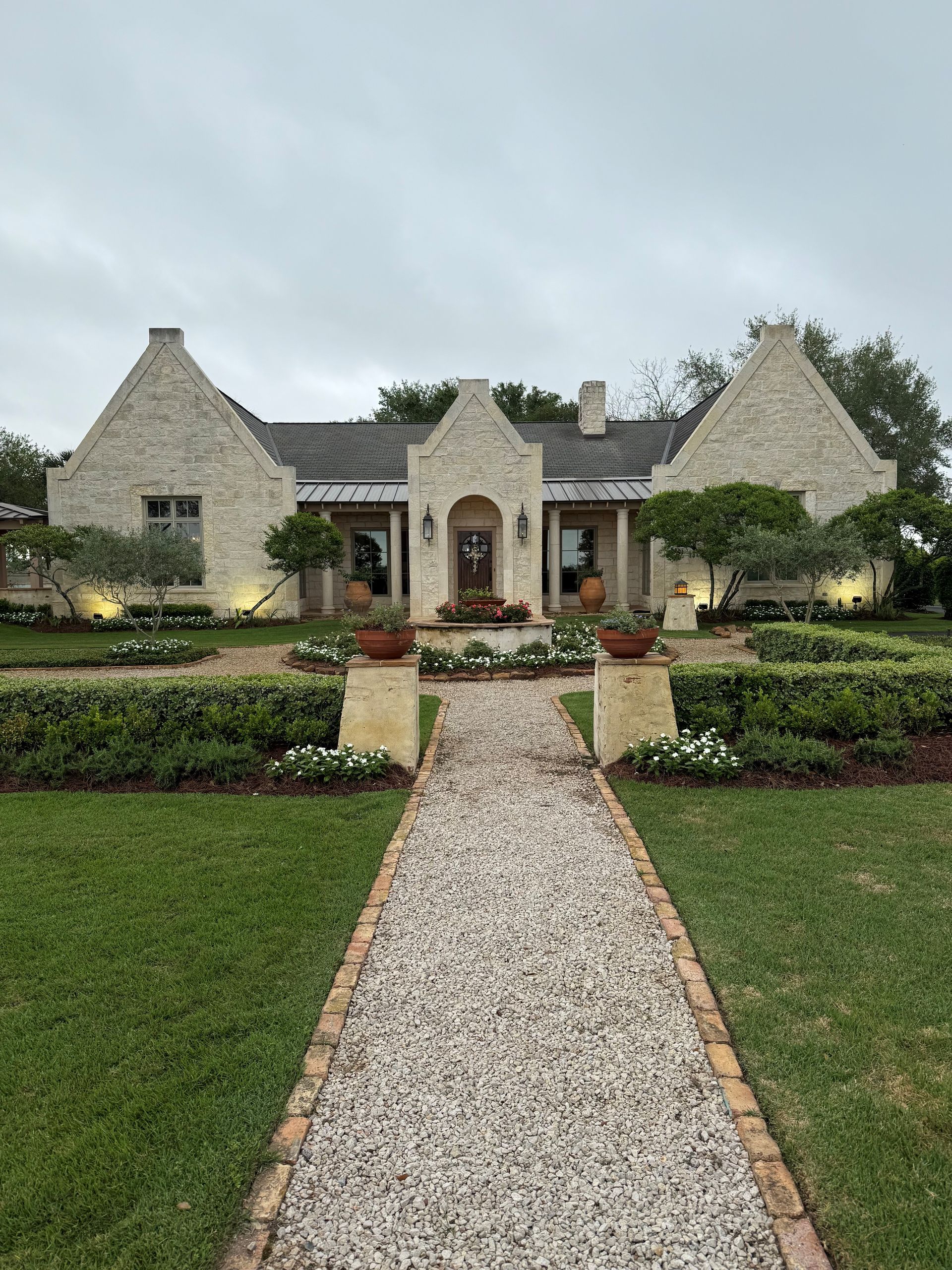 A beige stone cottage with a steep roof and peaked facade, approached by a long gravel path through a green lawn.