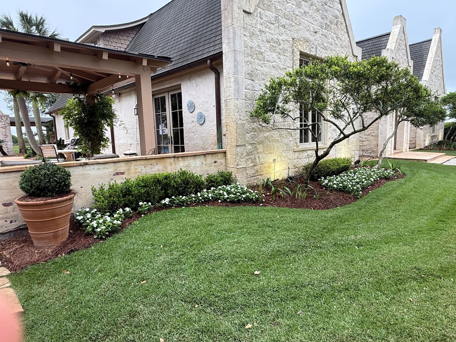 A stone cottage with a wooden pergola, manicured lawn, and landscaping featuring a small tree and white flower beds.
