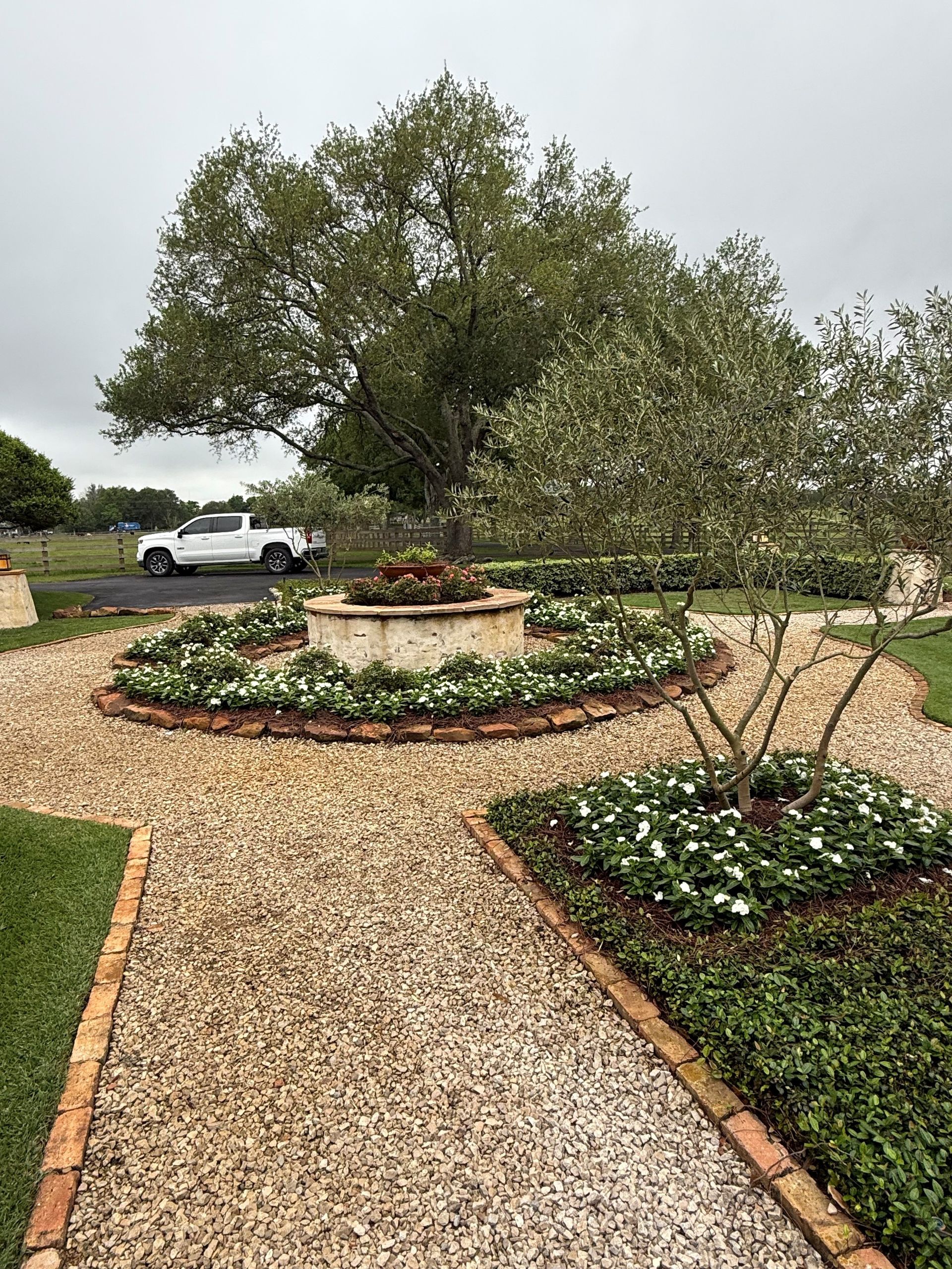 A gravel garden path leads to a central stone planter surrounded by flowering plants, with a white truck parked nearby.
