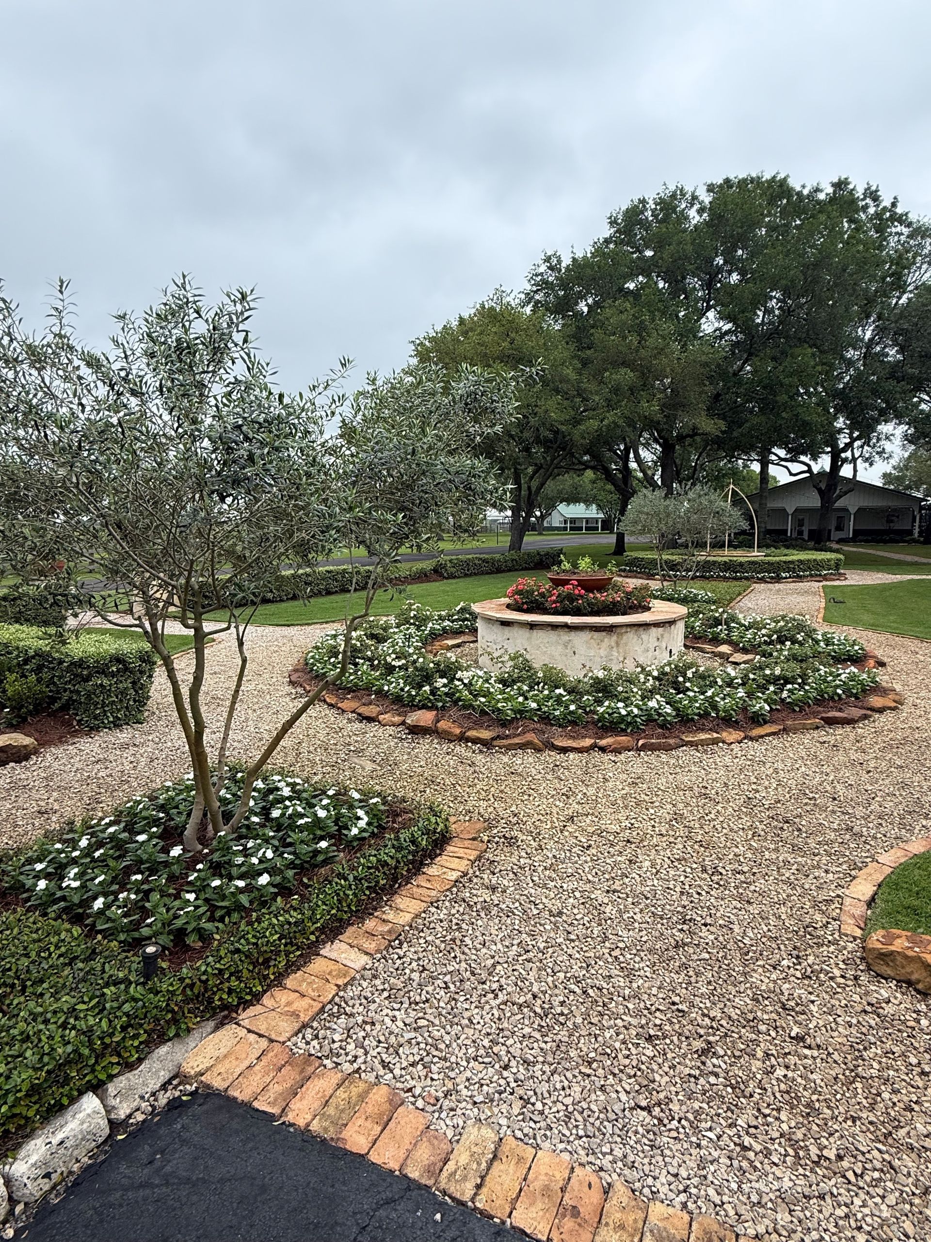 A gravel courtyard features a circular garden bed surrounding a central stone fountain, framed by trees and brick edging.