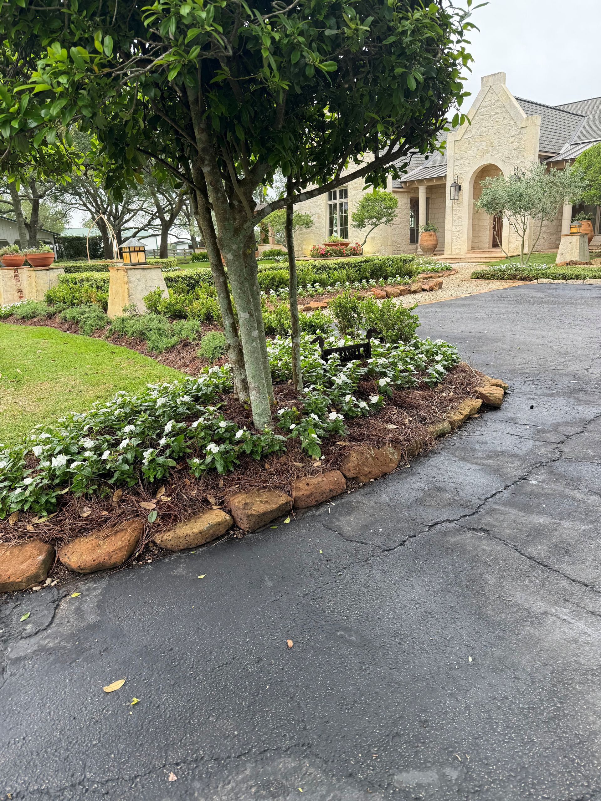 A light-colored stone cottage with a wooden pergola, manicured lawn, and landscaped flower beds under an overcast sky.