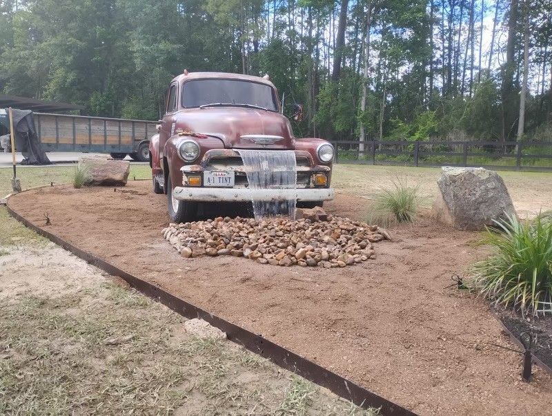 Rusty vintage truck with water feature, gravel bed, and boulders in a grassy outdoor setting.