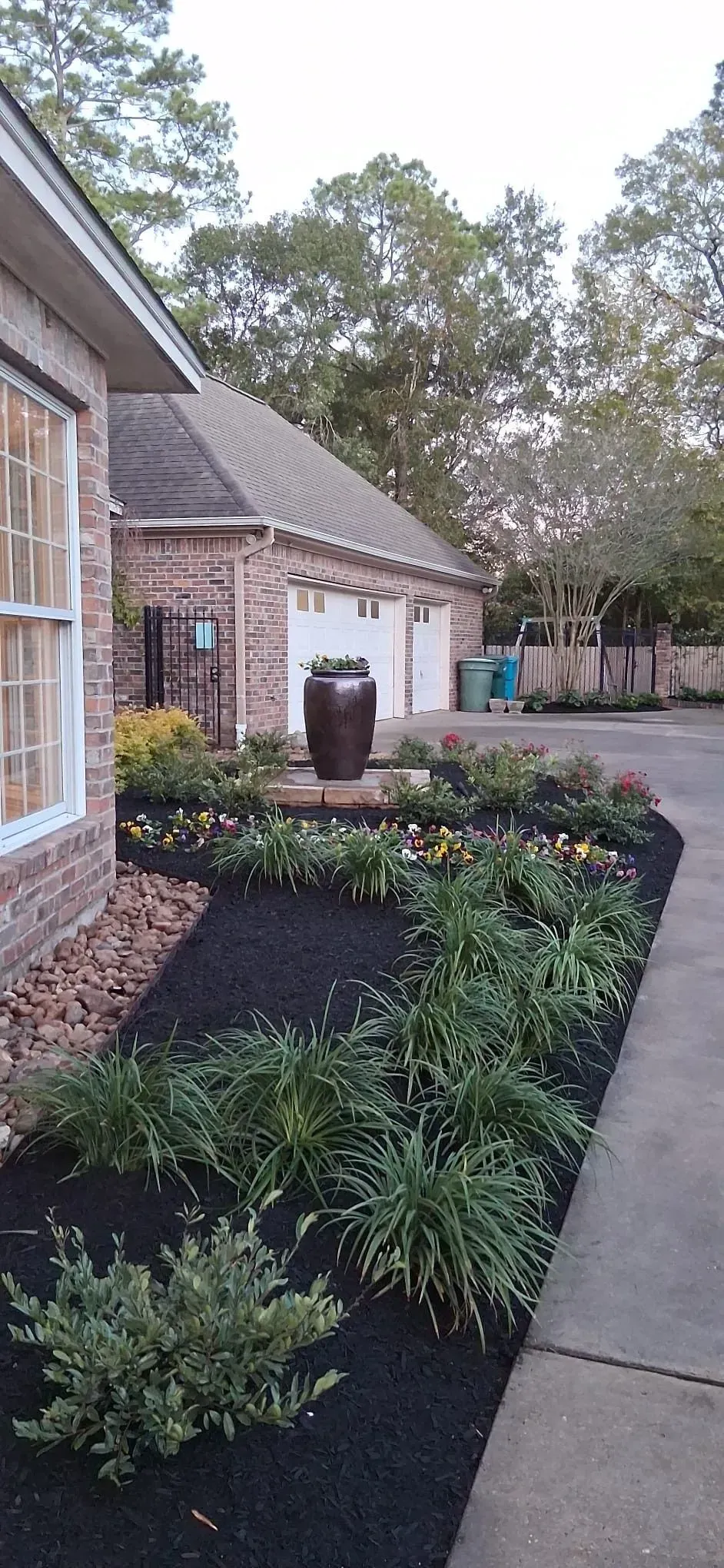 A well-landscaped garden with black mulch, green plants, and a dark brown vase in front of a brick house.