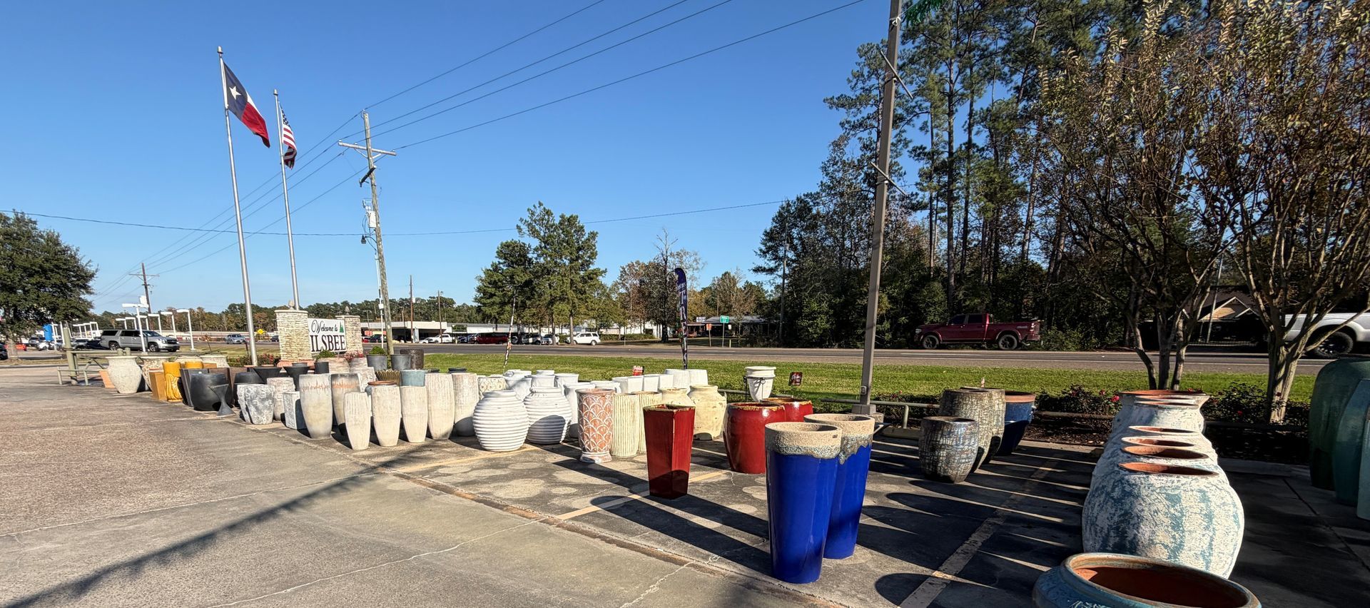 An outdoor display of various sized pottery in front of a Texas flag, next to a street with trees.