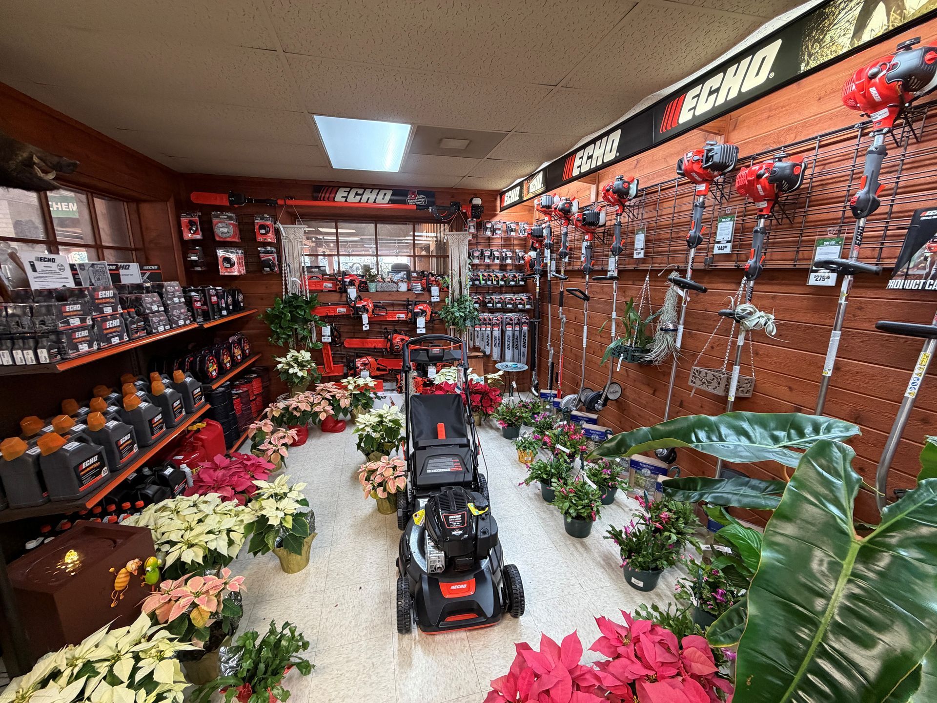 Inside a hardware store, a lawnmower sits surrounded by plants and tools, with displays of Echo brand equipment on walls.