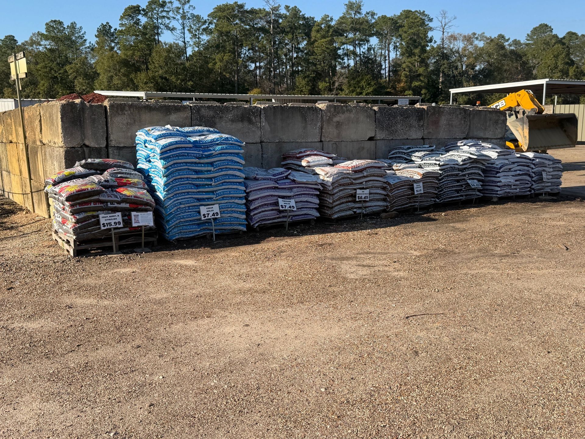 Bags of various colored landscaping materials for sale in an outdoor lot. Concrete blocks and a backhoe are visible.