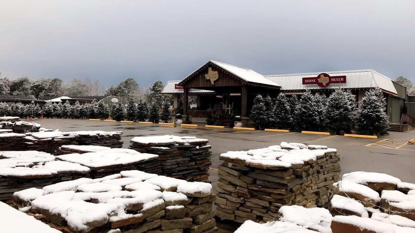 A snowy outdoor store with a brown wooden entrance, stacks of stone, and a row of evergreen trees.