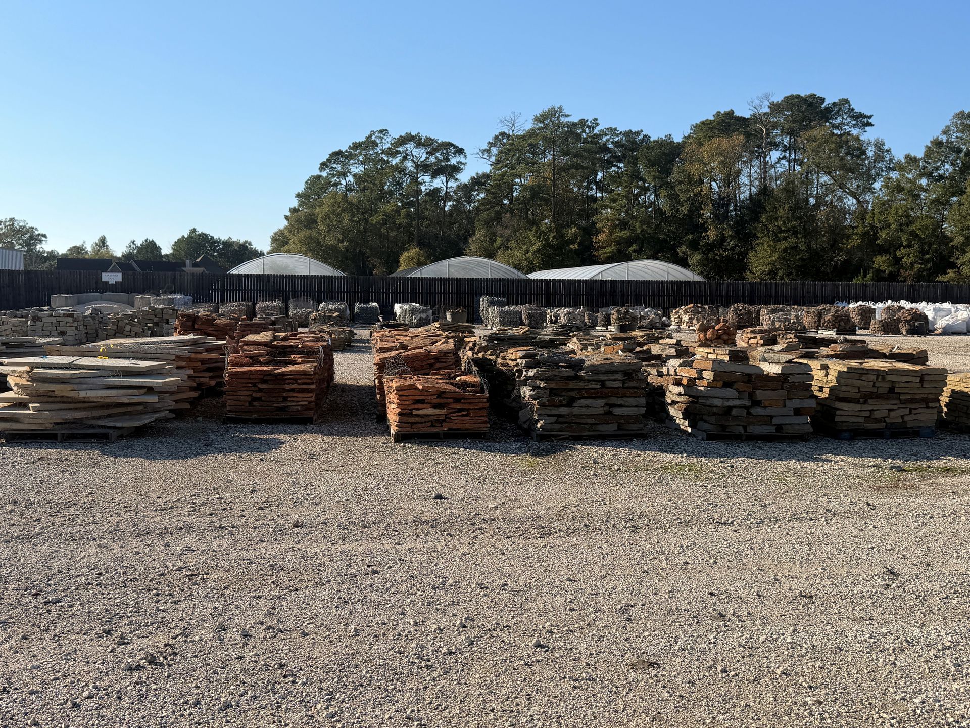 Stacks of various stone pavers, mostly tan and brown, in a gravel yard under a blue sky.