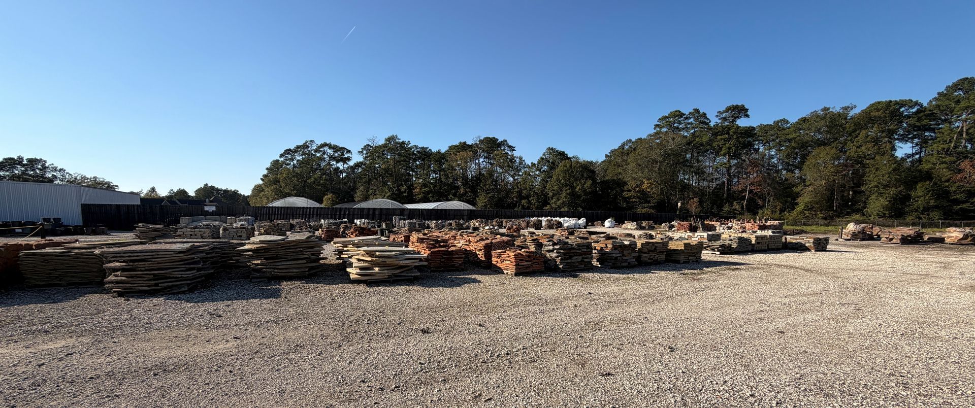A lumberyard with stacks of wooden planks, gravel ground, trees, and a clear blue sky.