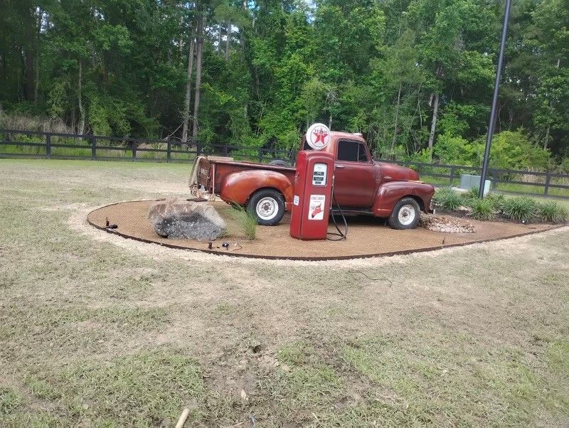 Red vintage truck and gas pump display on a circular gravel bed with a wooded backdrop.