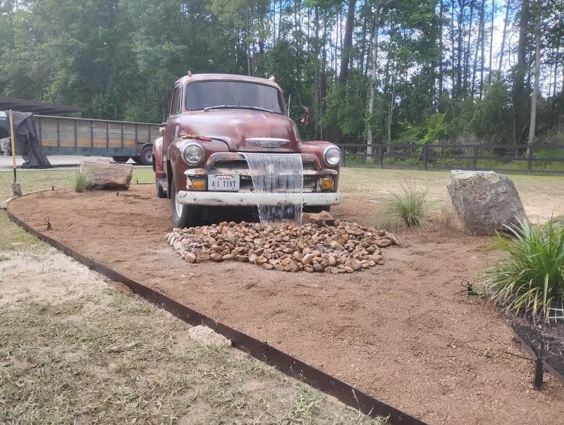 Old red truck with water cascading from its grill, surrounded by rocks and landscaping.