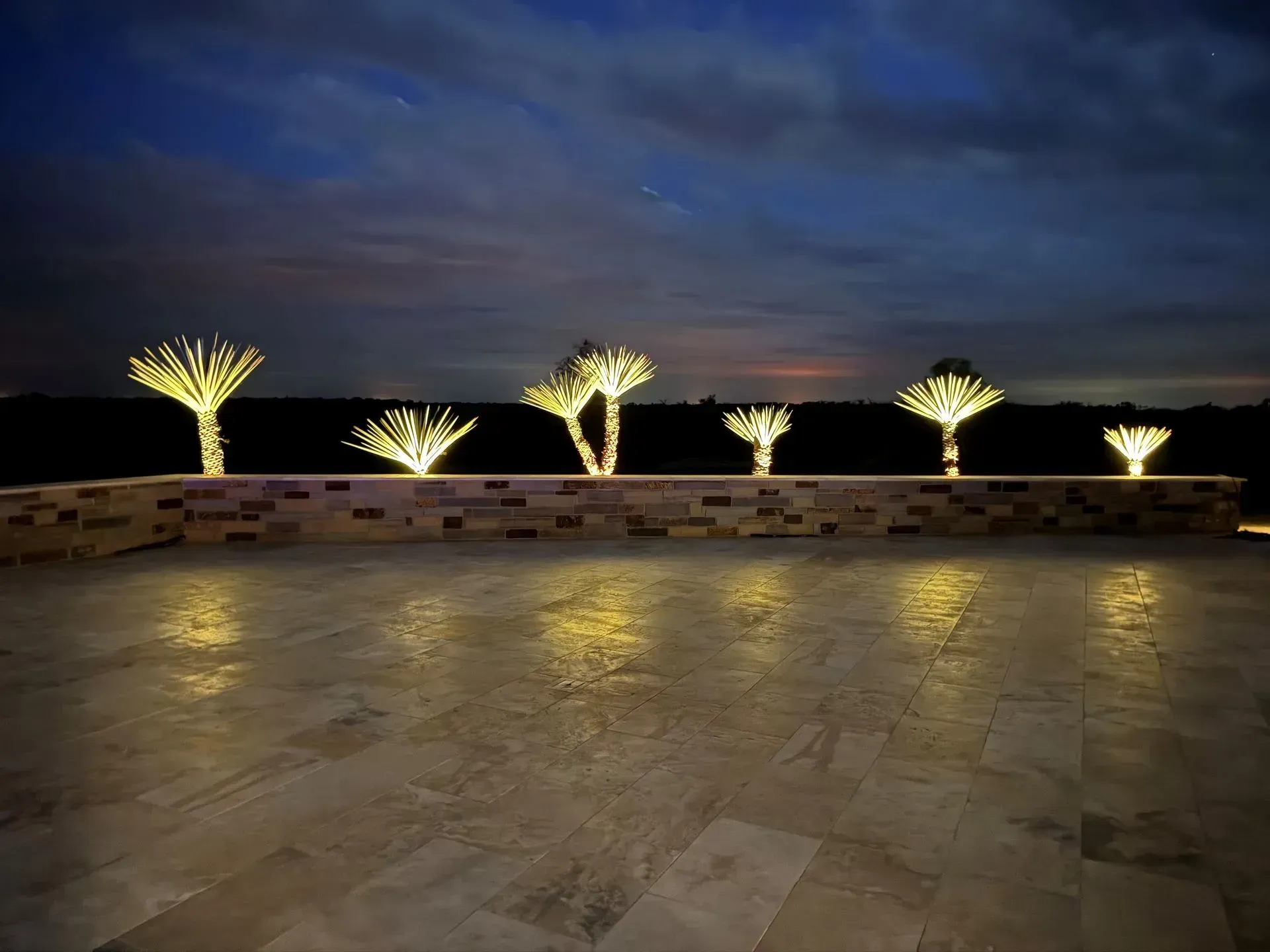 Night view: string lights illuminate small trees atop a brick wall; patio floor reflects their glow, dark sky.