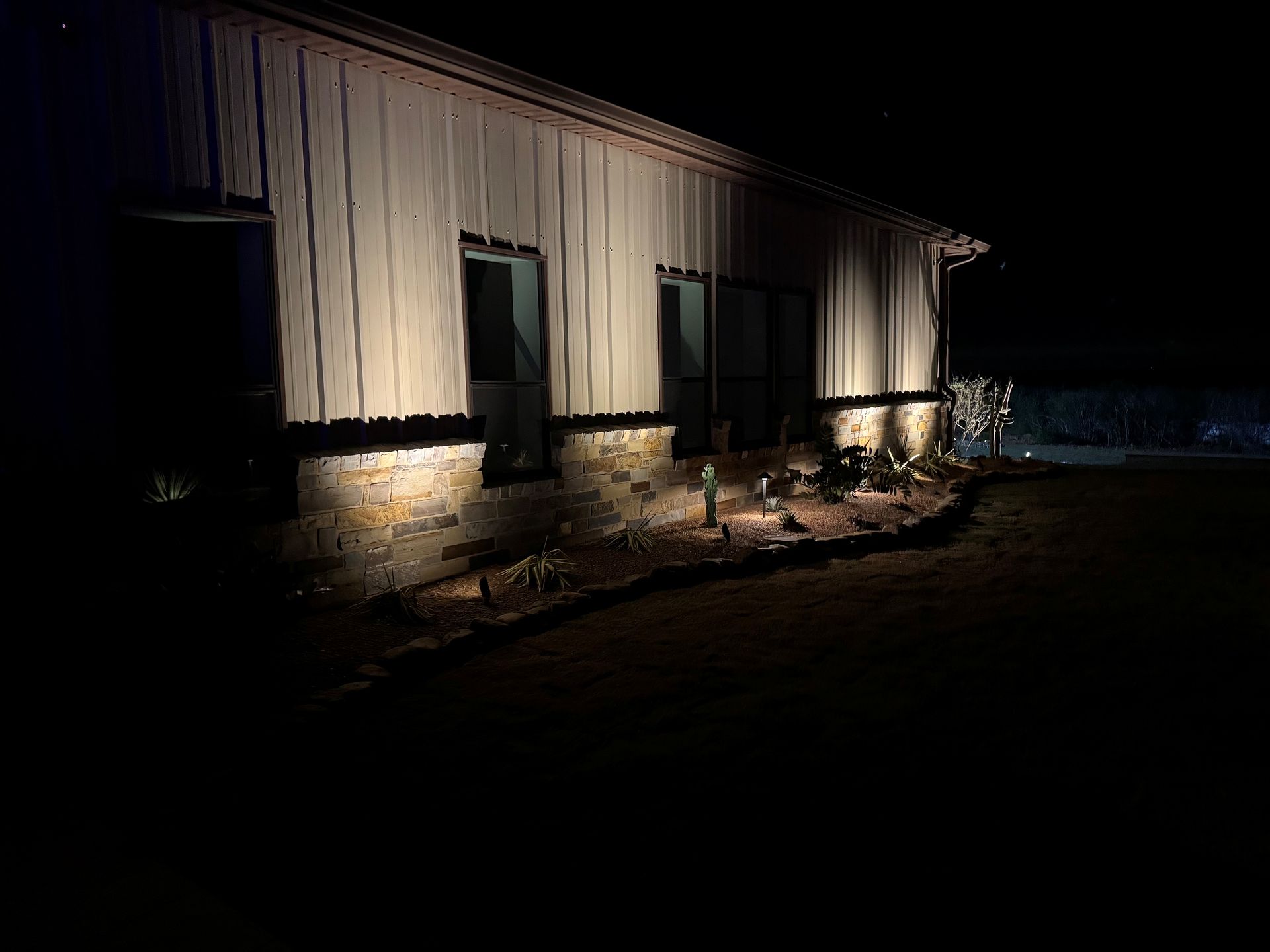 Night exterior view of a building illuminated by ground spotlights, highlighting its textured stone base and metal siding.