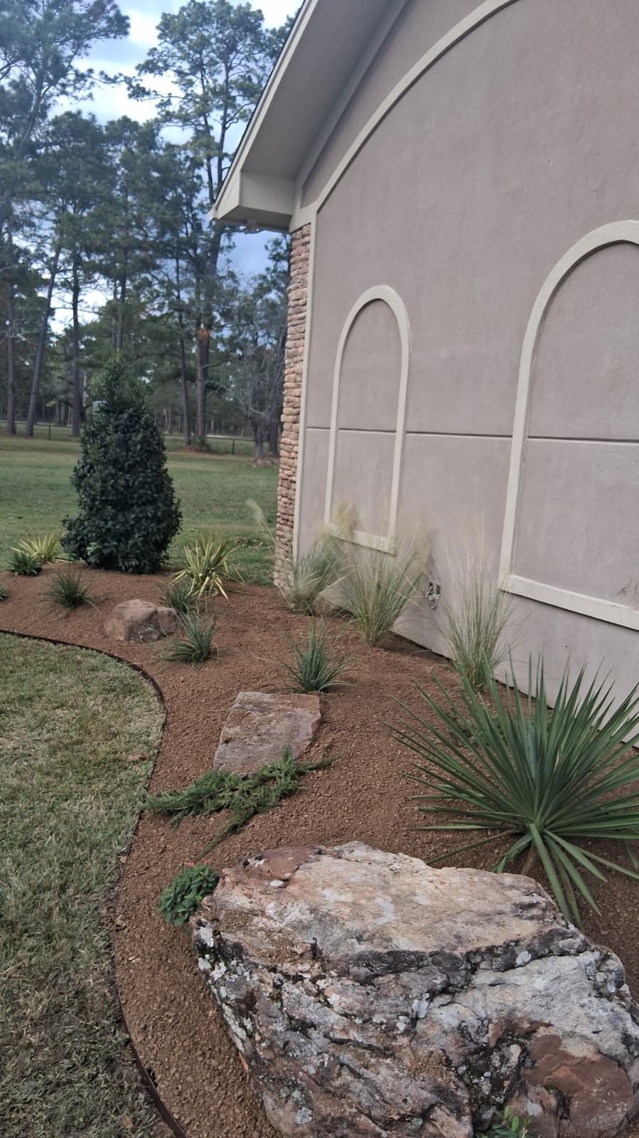 Landscaped flower bed with rocks and plants next to a tan stucco house and lawn.