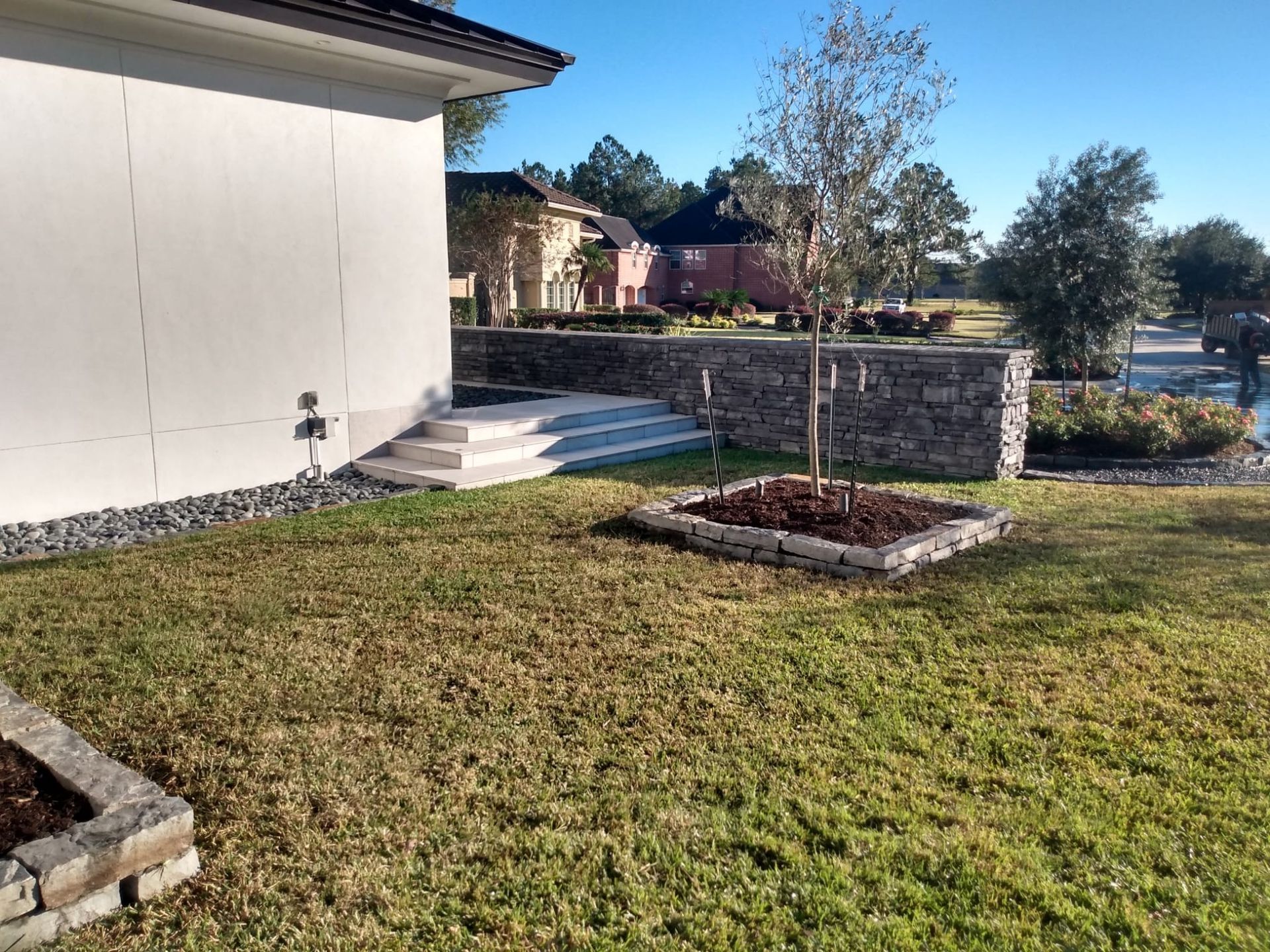 A modern house exterior with stone retaining walls, steps, and a young tree in a grassy yard.