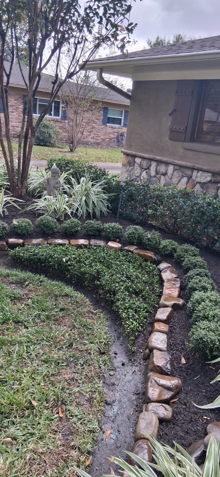 A landscaped garden bed with green plants, bordered by rocks, curves along a lawn in front of a house.