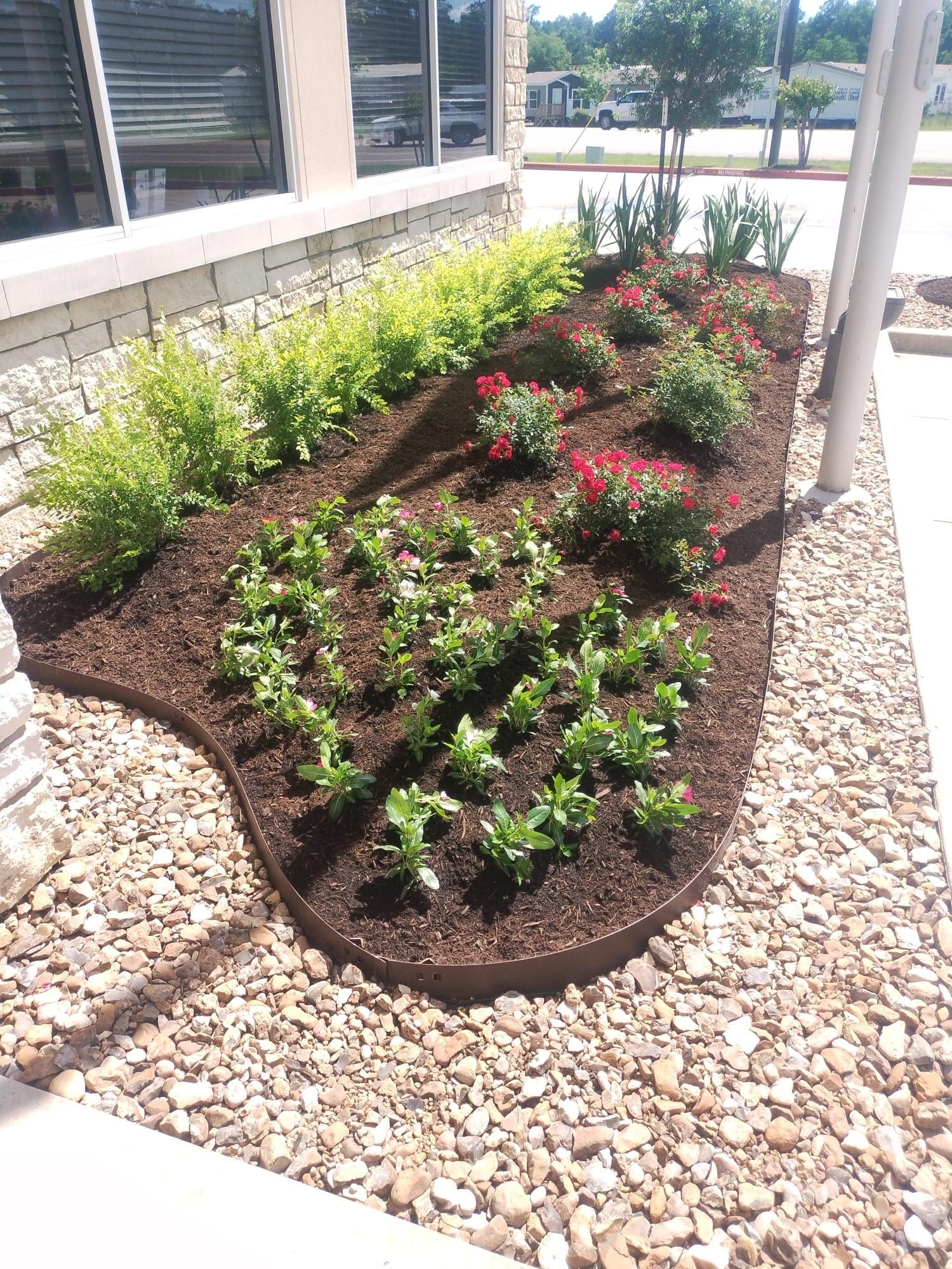 Flower bed with brown mulch, various green plants, red flowers, and river rock border.