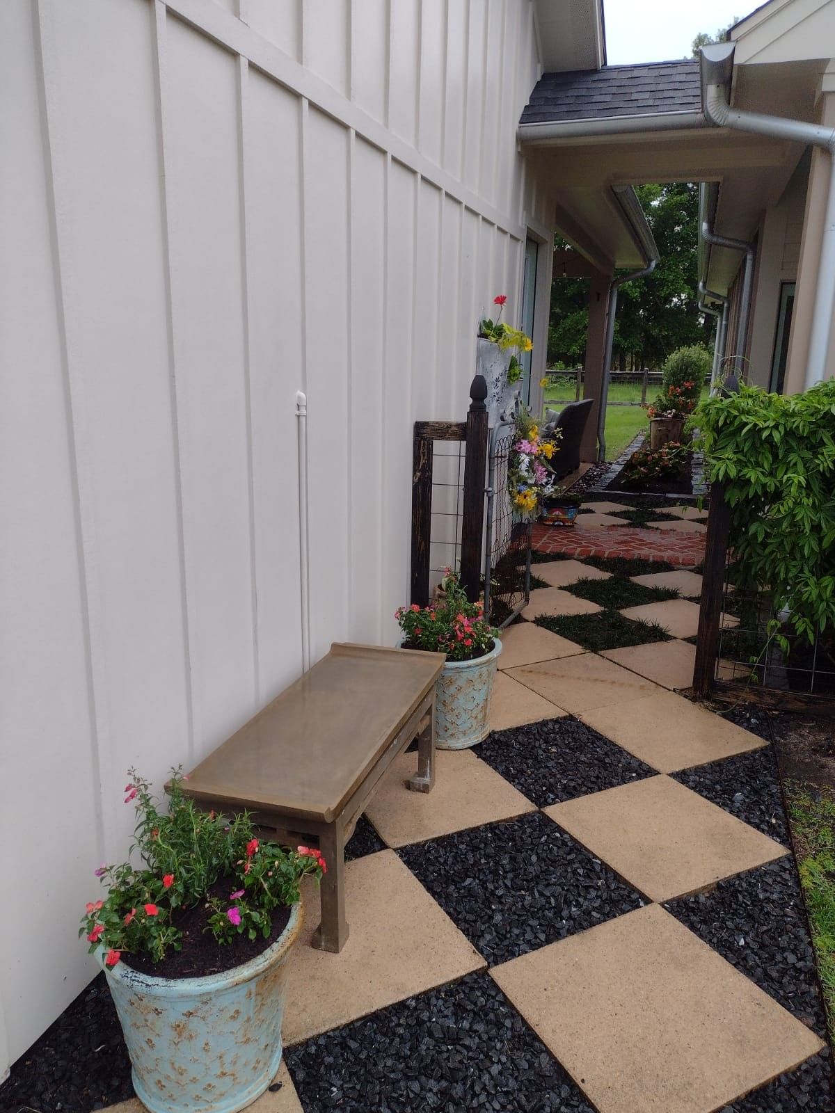 A bench sits beside a wall next to a checkered pathway. Planters with flowers line the path.