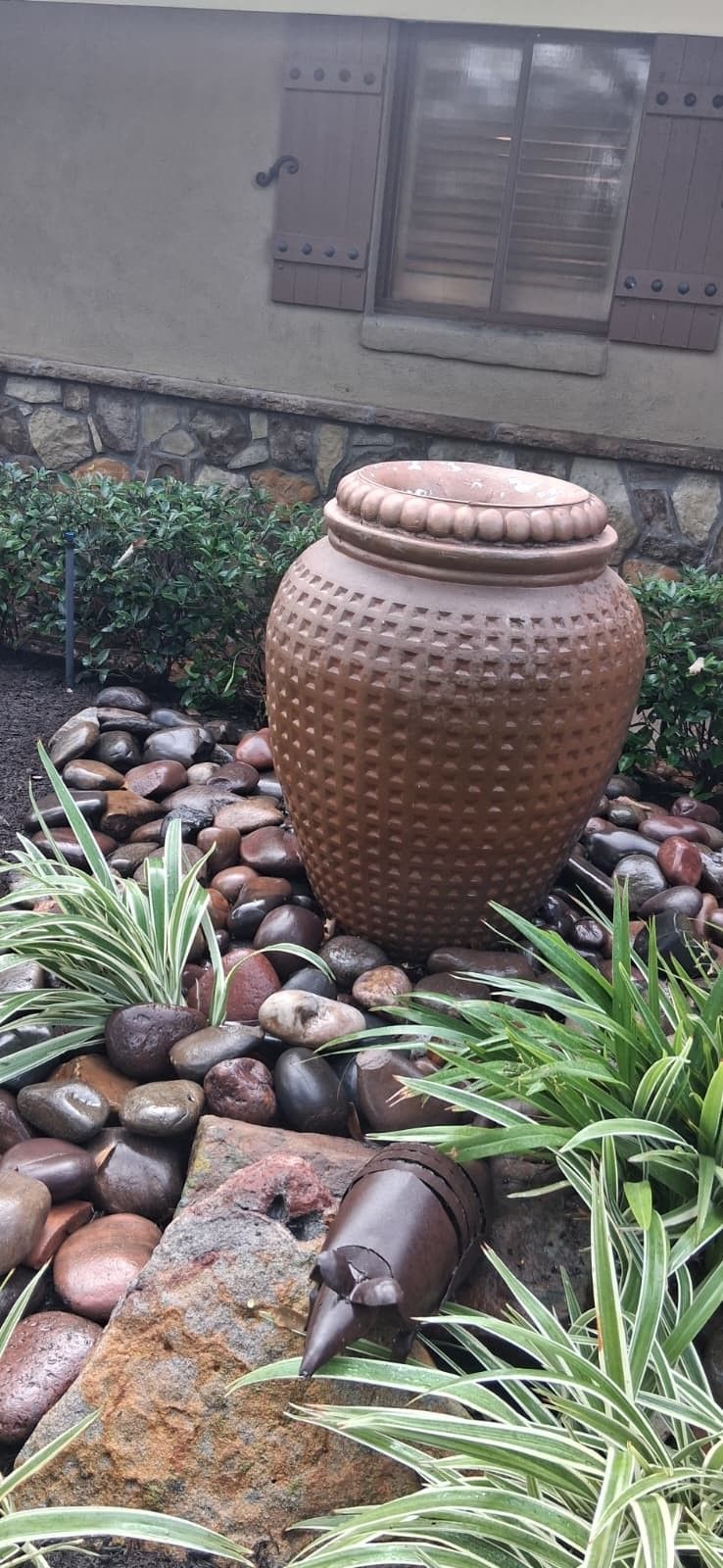 Brown ceramic jar fountain in a rock and plant garden by a building.