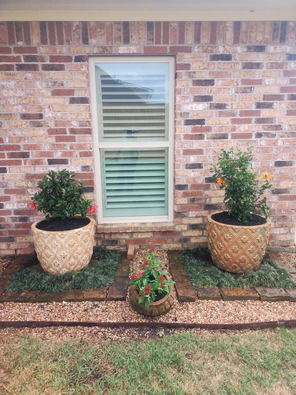Two large potted plants flank a window on a brick wall, with a smaller pot in front, all surrounded by gravel.