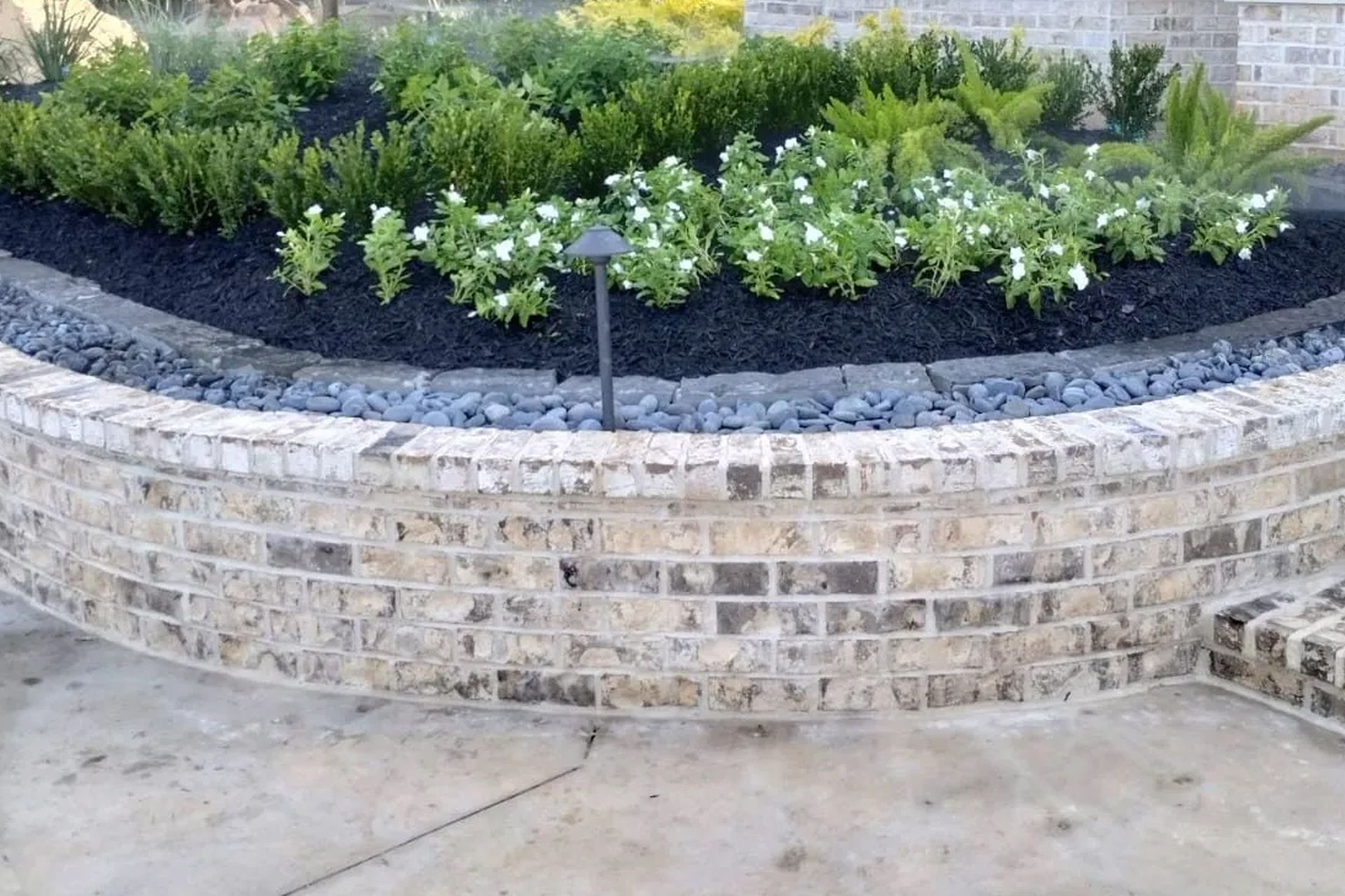 Brick retaining wall with landscaping. Green plants with white flowers, black mulch, and small black stones.